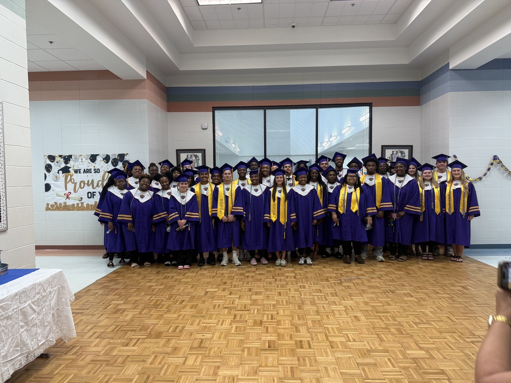 A group of graduating students in purple caps and gowns pose together in a classroom setting.