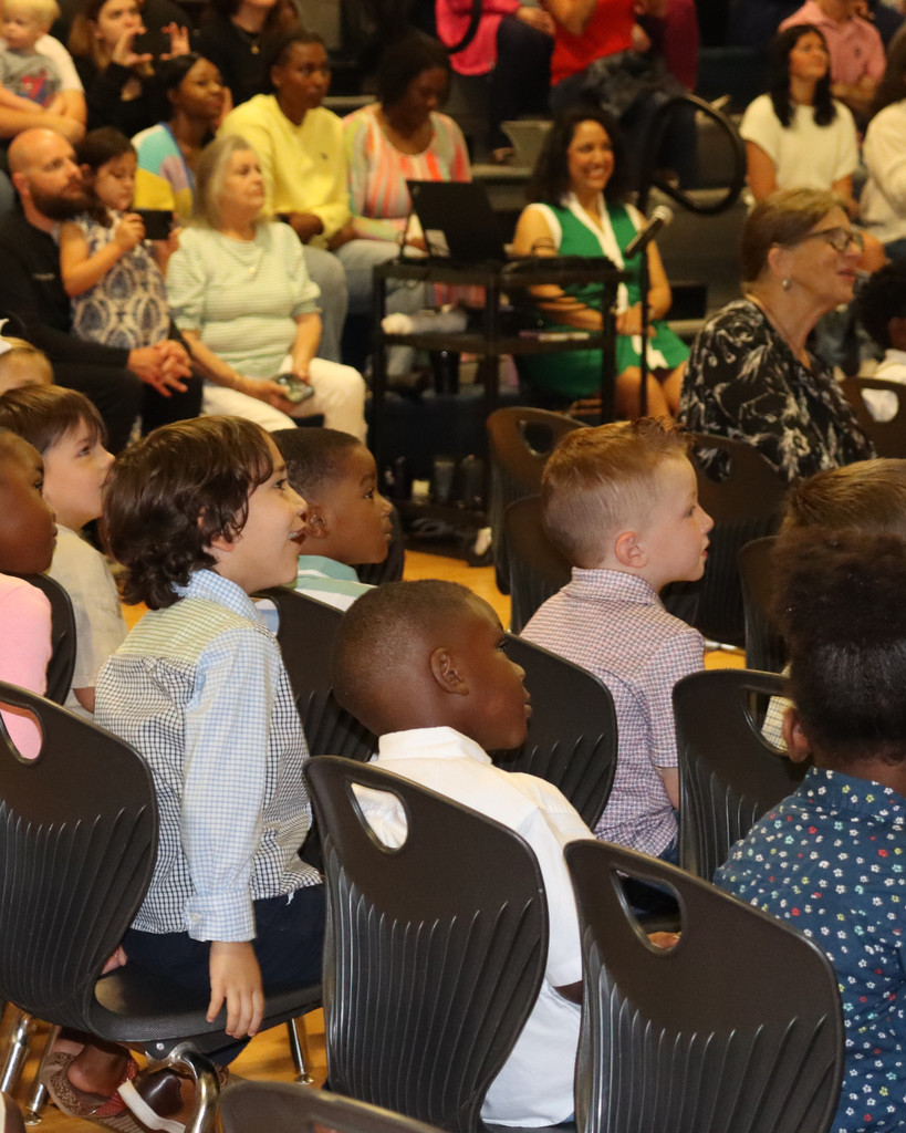 Pre-K students smile and watch the program while seated together in the school gymnasium.