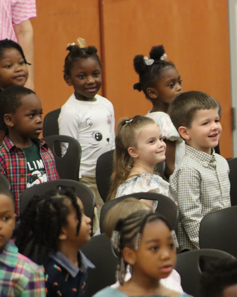 Pre-K students smile and watch the program while seated together in the school gymnasium.