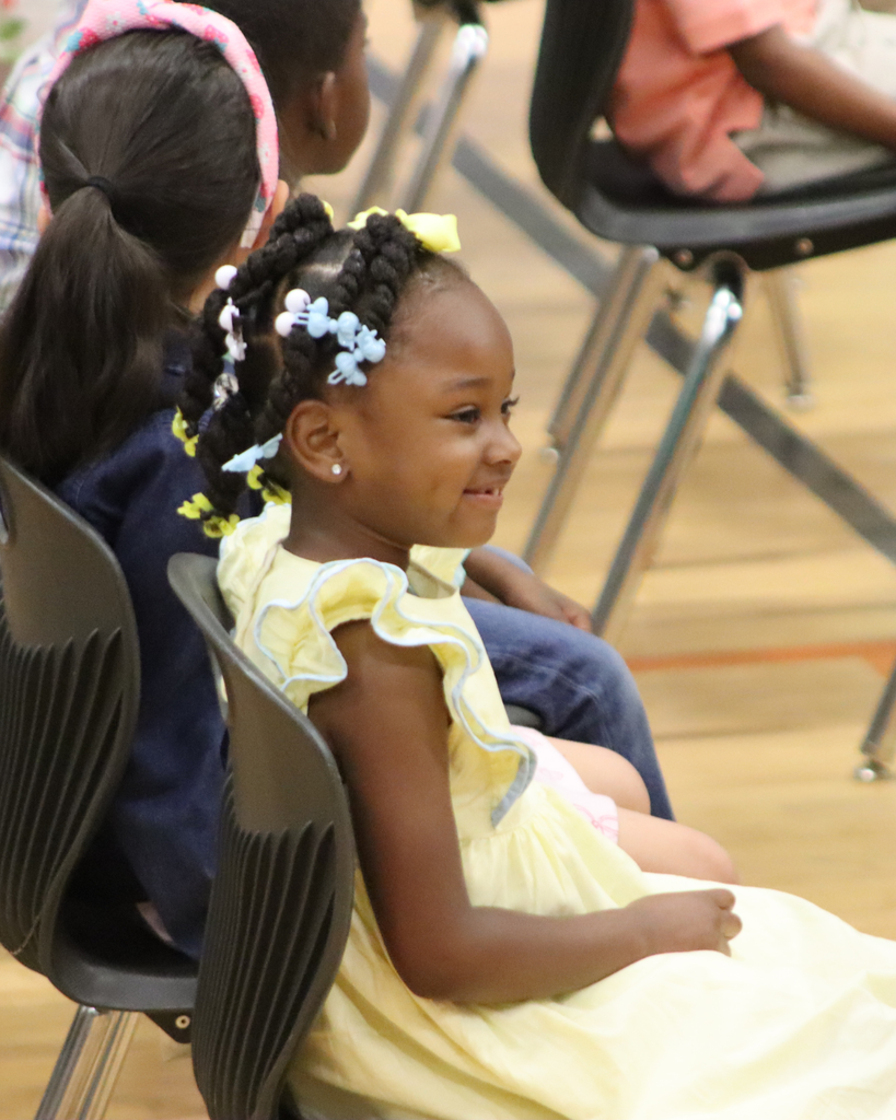 A smiling Pre-K student in a yellow dress sits with classmates during the JWP awards ceremony.