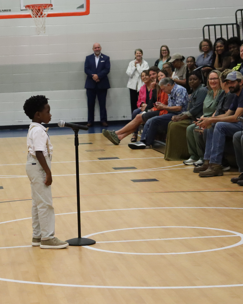 A Pre-K student stands at a microphone speaking during the JWP awards program while families and staff watch from the gymnasium seating area.