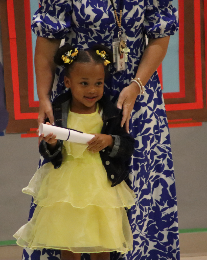 A young student smiles while holding a certificate during the JWP Pre-K awards ceremony with a teacher standing beside her.