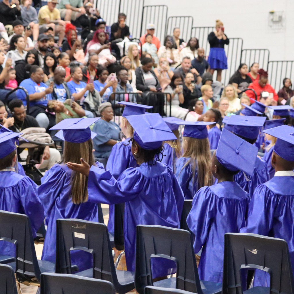 Kindergarten graduates in blue caps and gowns sit facing a crowd of families during a graduation ceremony.