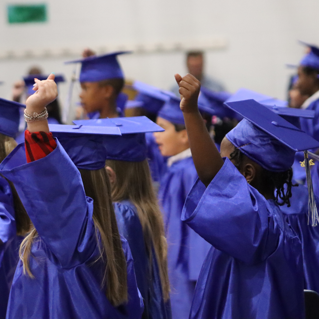 Young graduates in blue caps and gowns raise their hands during a school ceremony.