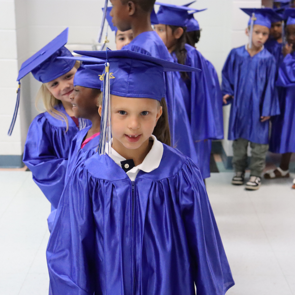 Smiling kindergarten graduate in a blue cap and gown looks back while seated with classmates.