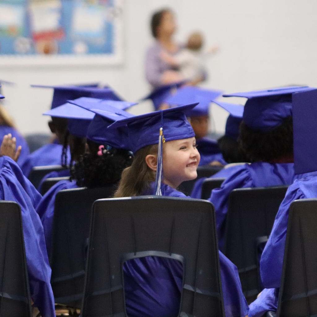 Smiling kindergarten graduate in a blue cap and gown looks back while seated with classmates.