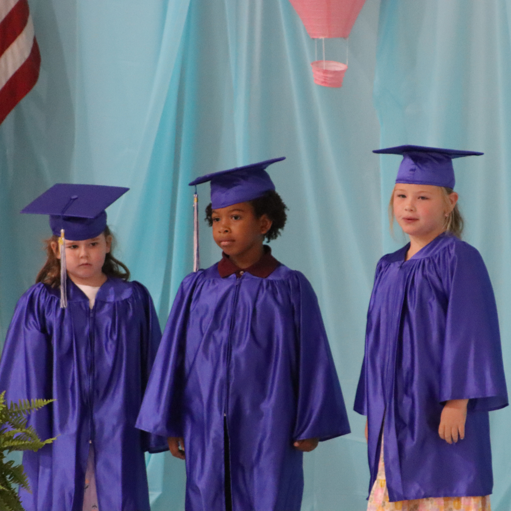Three kindergarten graduates stand on stage in blue caps and gowns during graduation.