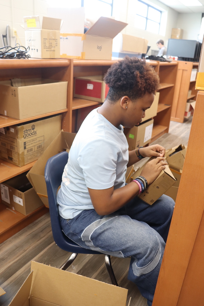 Student assembling or folding a cardboard box while seated in a workspace with shelves and boxes.