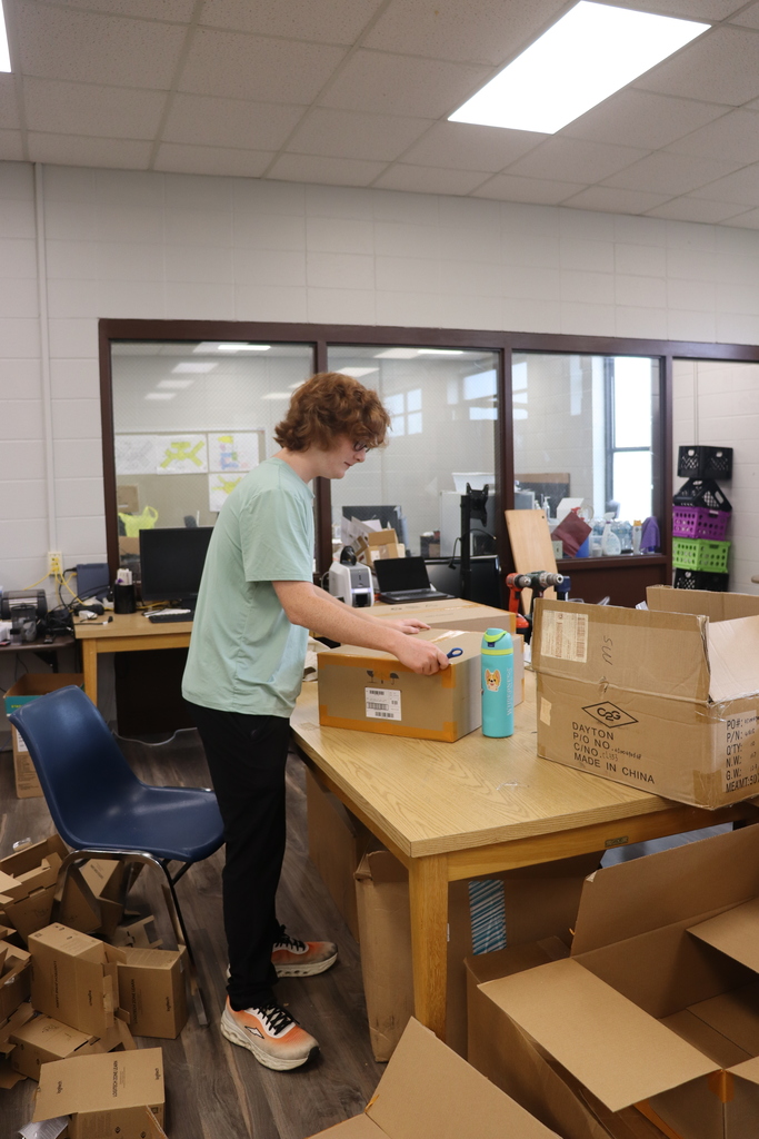Student opening a cardboard box on a worktable surrounded by equipment and packaging materials.