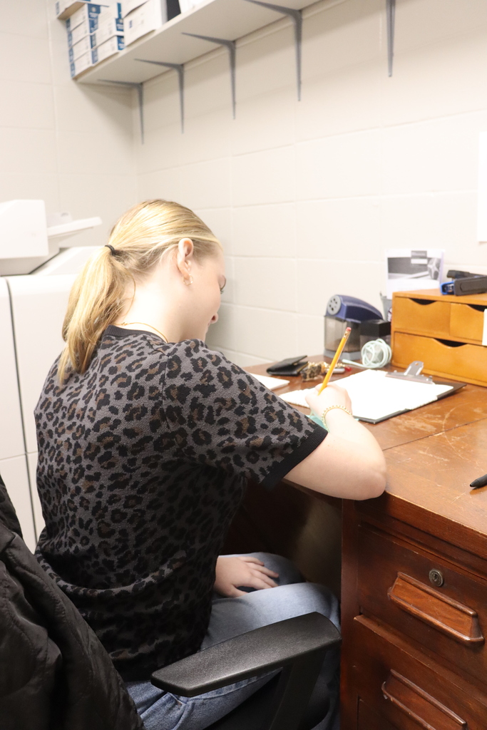 : Student seated at a desk, writing on a clipboard in an office workspace.