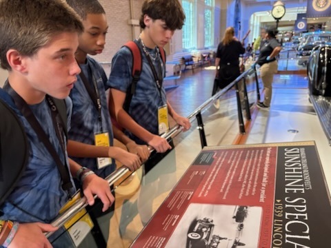 A series of photos shows three middle school students participating in a STEM and innovation event at a museum. The students explore interactive exhibits, including a flight simulator and historical automobile displays, while learning about engineering and technology. They examine informational exhibits, speak with event organizers and judges about STEM projects, and view a prototype backpack designed with ventilation and cooling features. Additional photos show the students receiving event materials, engaging in discussions about their project, and posing outdoors beside a large Abraham Lincoln mascot figure. The images capture hands-on learning, teamwork, problem-solving, and student engagement in science, technology, engineering, and innovation activities.