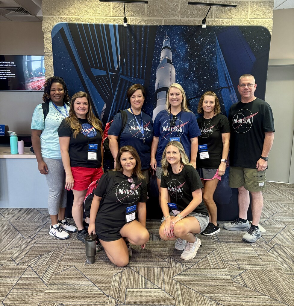 A group of educators wearing NASA shirts pose in front of a Saturn V rocket display during a training visit.