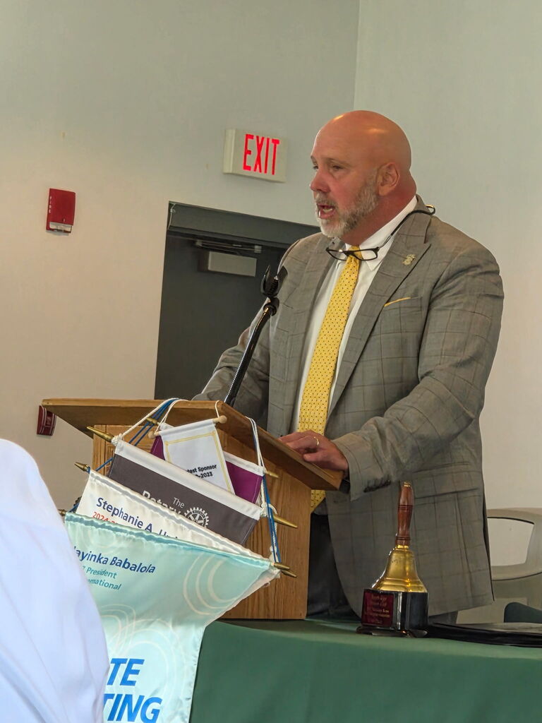 Superintendent Dr. Boyd English speaks at a podium during a Bainbridge Rotary Club meeting. Rotary banners and a brass bell are displayed in front of him.
