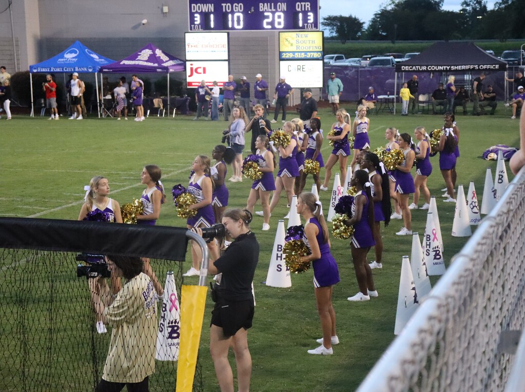 Bainbridge High School cheerleaders stand along the football sideline with pom-poms.