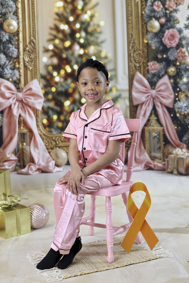 A young girl wearing pink satin pajamas and a black headband sits smiling on a pink chair in front of a festive Christmas backdrop decorated with pink bows, ornaments, and lights. A gold childhood cancer awareness ribbon is displayed beside her.