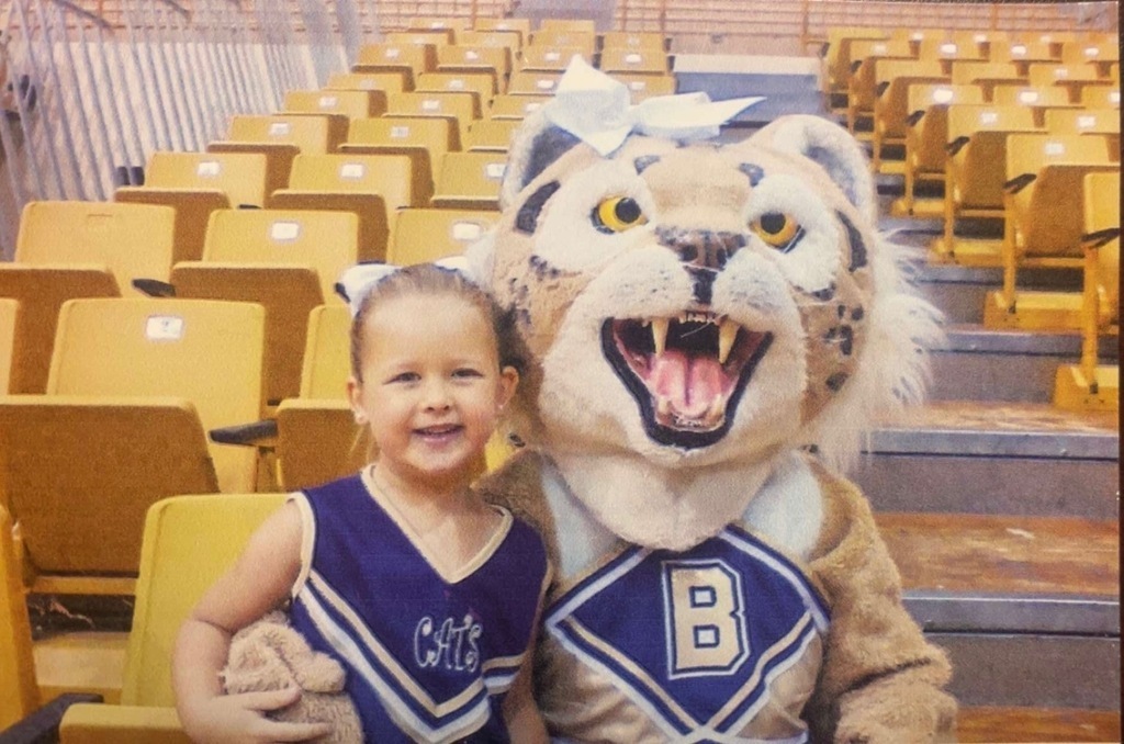 create short alt text  Young cheerleader posing with a Bearcat mascot in a school gym.