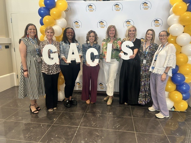 Seven women pose together holding letters spelling “GACIS” in front of a GACIS backdrop with blue, gold, and white balloons.