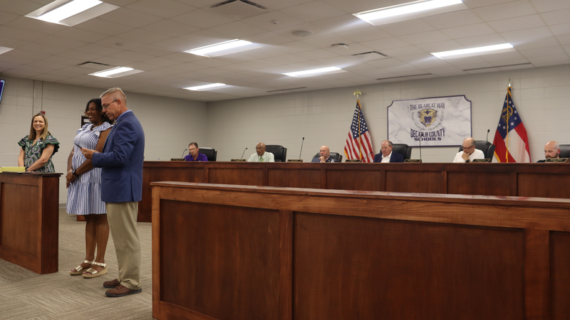 The image shows a school board meeting with three people standing in front presenting or recognizing someone. Five men sit behind a wooden dais with an American flag and a state flag. A sign above reads: "THE BEARCAT WAY DECATUR COUNTY SCHOOLS."