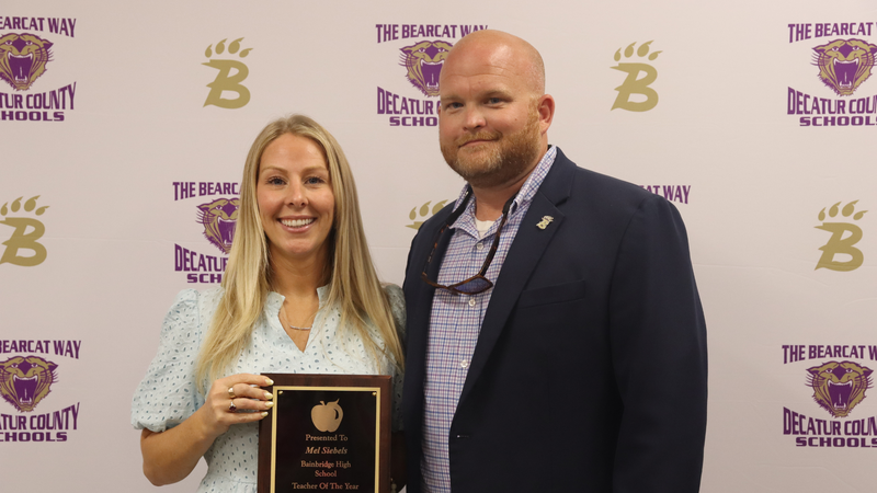 Teacher and principal stand holding award in front of a branded decatur county schools media backdrop