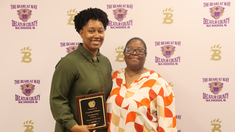 Teacher and principal stand holding award in front of a branded decatur county schools media backdrop