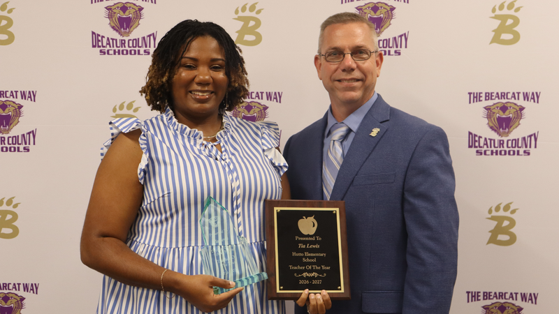 Teacher and principal stand holding award in front of a branded decatur county schools media backdrop