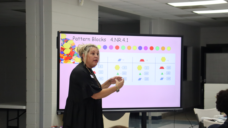 A presenter gestures while explaining a math lesson displayed on a large screen titled “Pattern Blocks 4.NR.4.1,” which shows colorful geometric shapes and fractions.