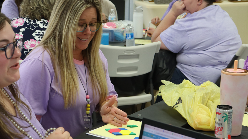 A woman in a lavender shirt smiles while arranging colorful pattern blocks on a yellow board during a group learning activity.
