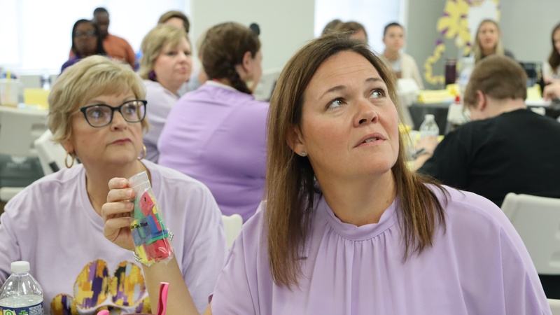 Two women wearing light purple shirts sit at a table during a group workshop or meeting, one holding a small plastic bag with colorful items while looking upward attentively.