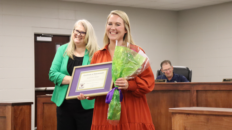 A smiling award recipient holds a framed certificate and bouquet while standing beside another woman during a recognition at a board meeting.