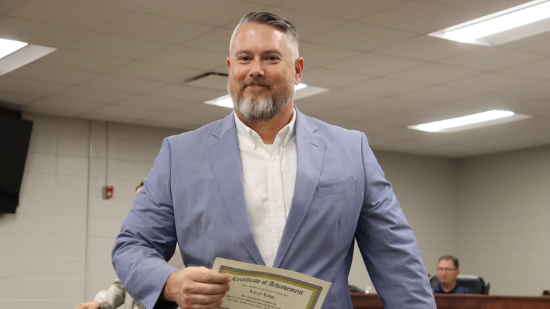 A man in a light blue blazer smiles while holding a certificate during a recognition at a board meeting.