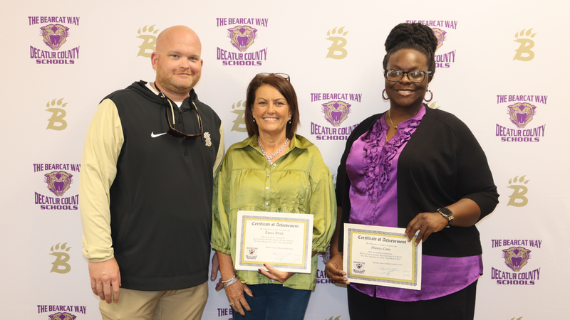 Three individuals stand in front of a Decatur County Schools backdrop, with two holding certificates of achievement and smiling.