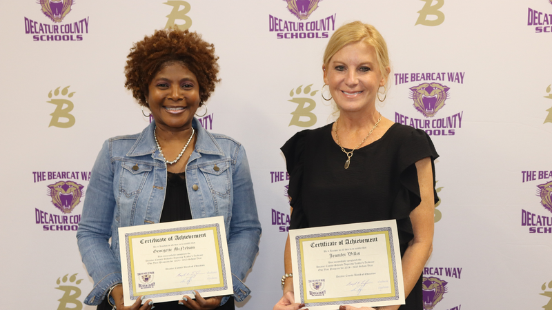 Two women stand in front of a Decatur County Schools backdrop, each holding a certificate of achievement and smiling.