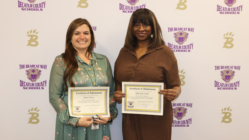 Two women stand in front of a Decatur County Schools backdrop, each holding a certificate of achievement and smiling.