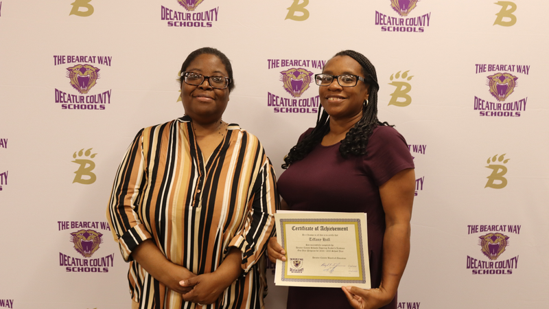 Two women stand in front of a Decatur County Schools backdrop, each holding a certificate of achievement and smiling.