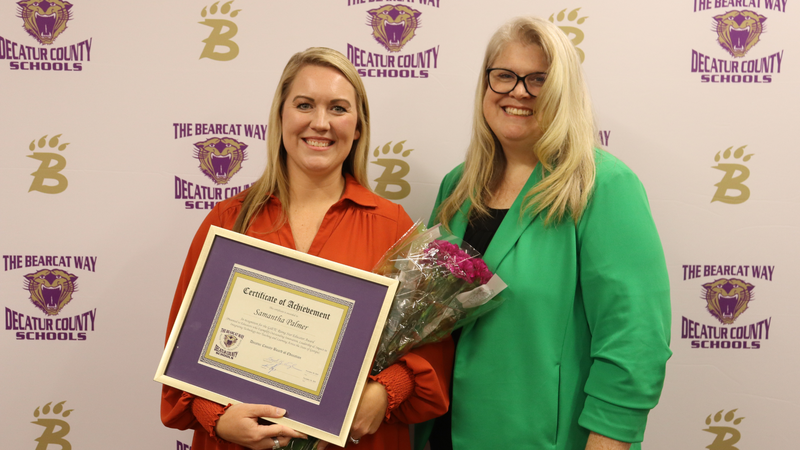 Two women stand in front of a Decatur County Schools backdrop; one holds a framed certificate and bouquet while both smile.