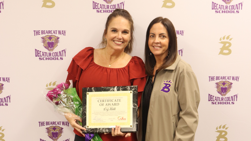 Two women stand in front of a Decatur County Schools backdrop; one holds a framed certificate and bouquet while both smile.