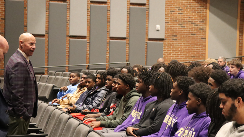 Coach stands in the aisle speaking closely with seated students in an auditorium.