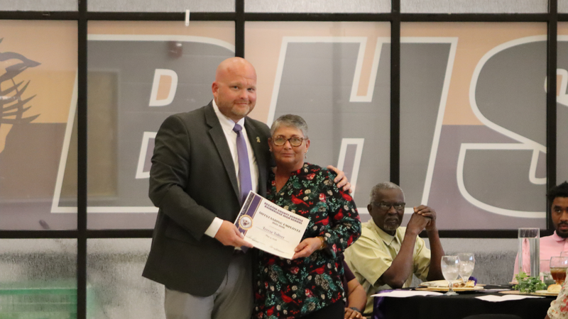 An award recipient poses with a presenter while holding a certificate at a recognition event, with attendees seated at tables in the background.