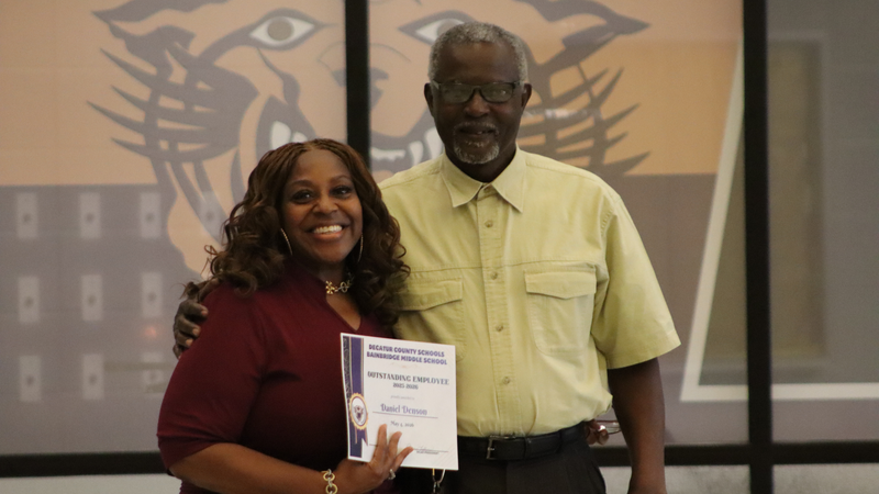 An award recipient poses with a presenter while holding a certificate at a recognition event, with attendees seated at tables in the background.