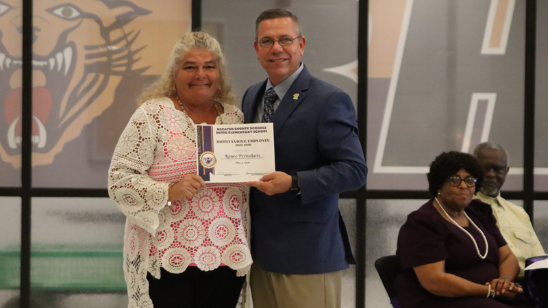 An award recipient poses with a presenter while holding a certificate at a recognition event, with attendees seated at tables in the background.