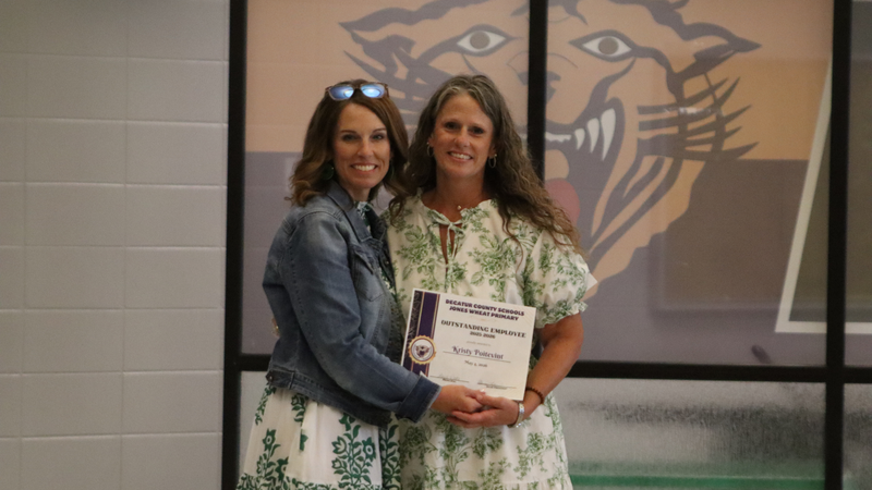 An award recipient poses with a presenter while holding a certificate at a recognition event, with attendees seated at tables in the background.