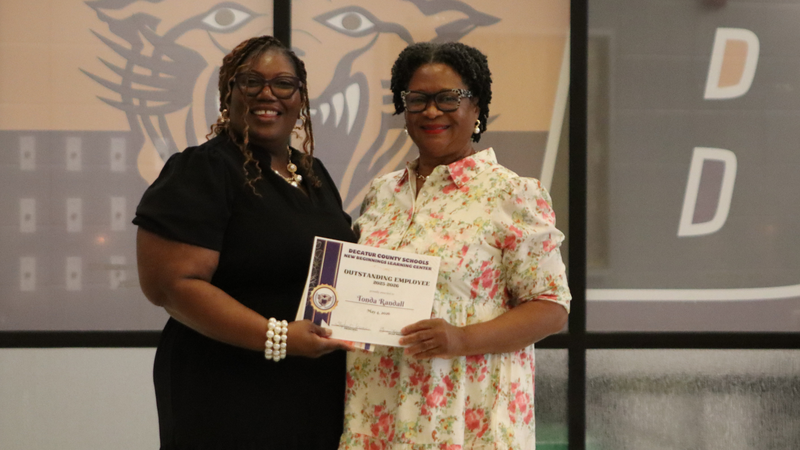 An award recipient poses with a presenter while holding a certificate at a recognition event, with attendees seated at tables in the background.