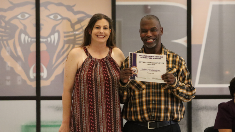 An award recipient poses with a presenter while holding a certificate at a recognition event, with attendees seated at tables in the background.
