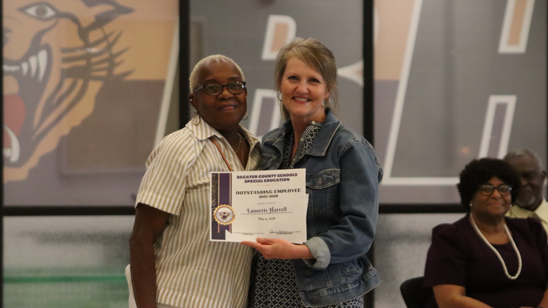 An award recipient poses with a presenter while holding a certificate at a recognition event, with attendees seated at tables in the background.