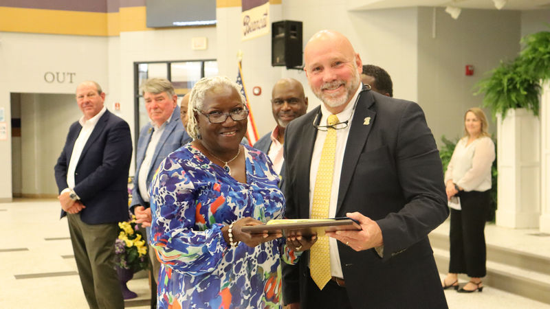 An award recipient poses with a presenter while holding a certificate at a recognition event, with attendees seated at tables in the background.