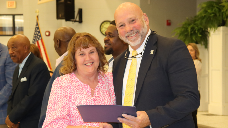 An award recipient poses with a presenter while holding a certificate at a recognition event, with attendees seated at tables in the background.