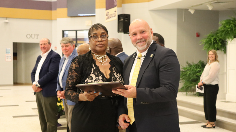 An award recipient poses with a presenter while holding a certificate at a recognition event, with attendees seated at tables in the background.