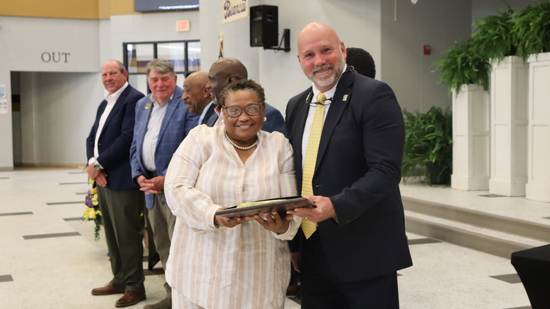 An award recipient poses with a presenter while holding a certificate at a recognition event, with attendees seated at tables in the background.