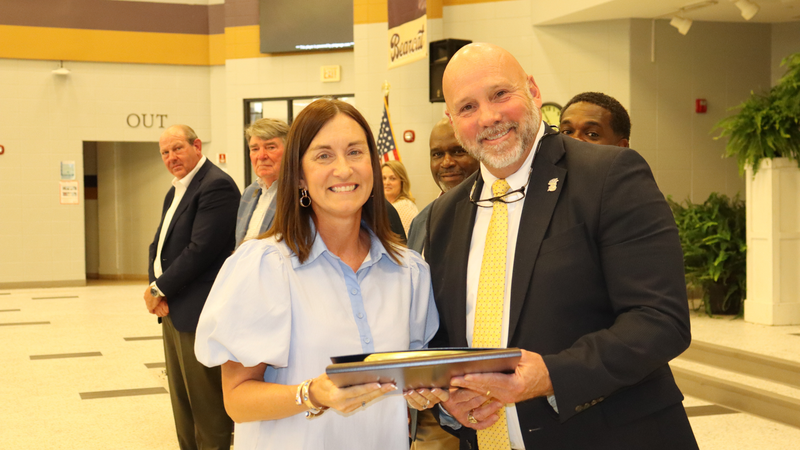 An award recipient poses with a presenter while holding a certificate at a recognition event, with attendees seated at tables in the background.
