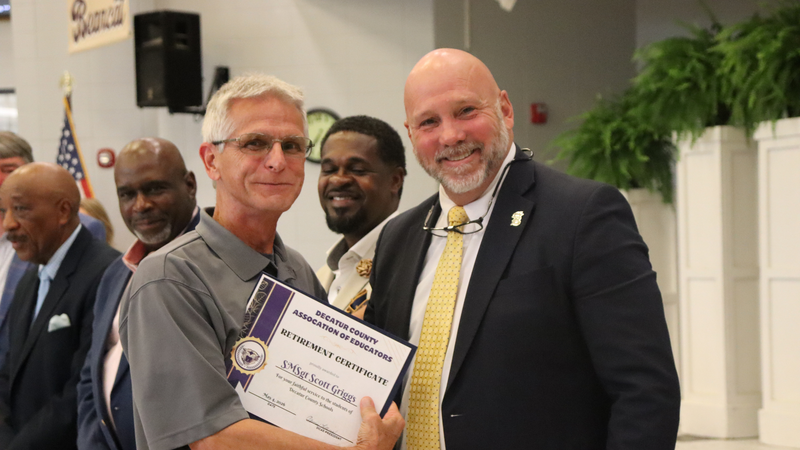 An award recipient poses with a presenter while holding a certificate at a recognition event, with attendees seated at tables in the background.
